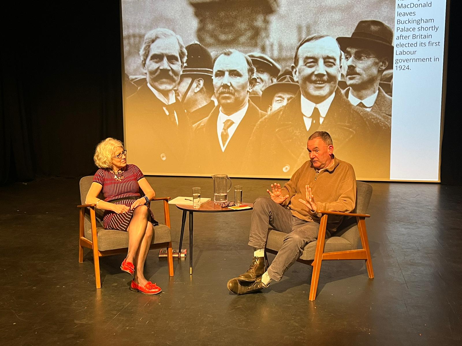 A woman interviewer, Liz Jerrard, seated on a stage with Jon Cruddas MP with a photographic backdrop of Ramsey MacDonald and comrades.